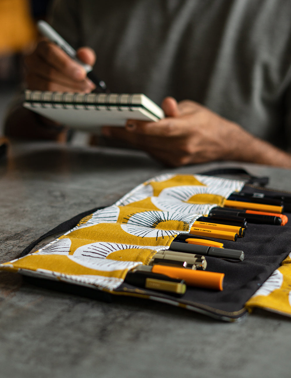 An artist sketching in a cafe with a beautiful roll up case filled with art supplies, open next to them.