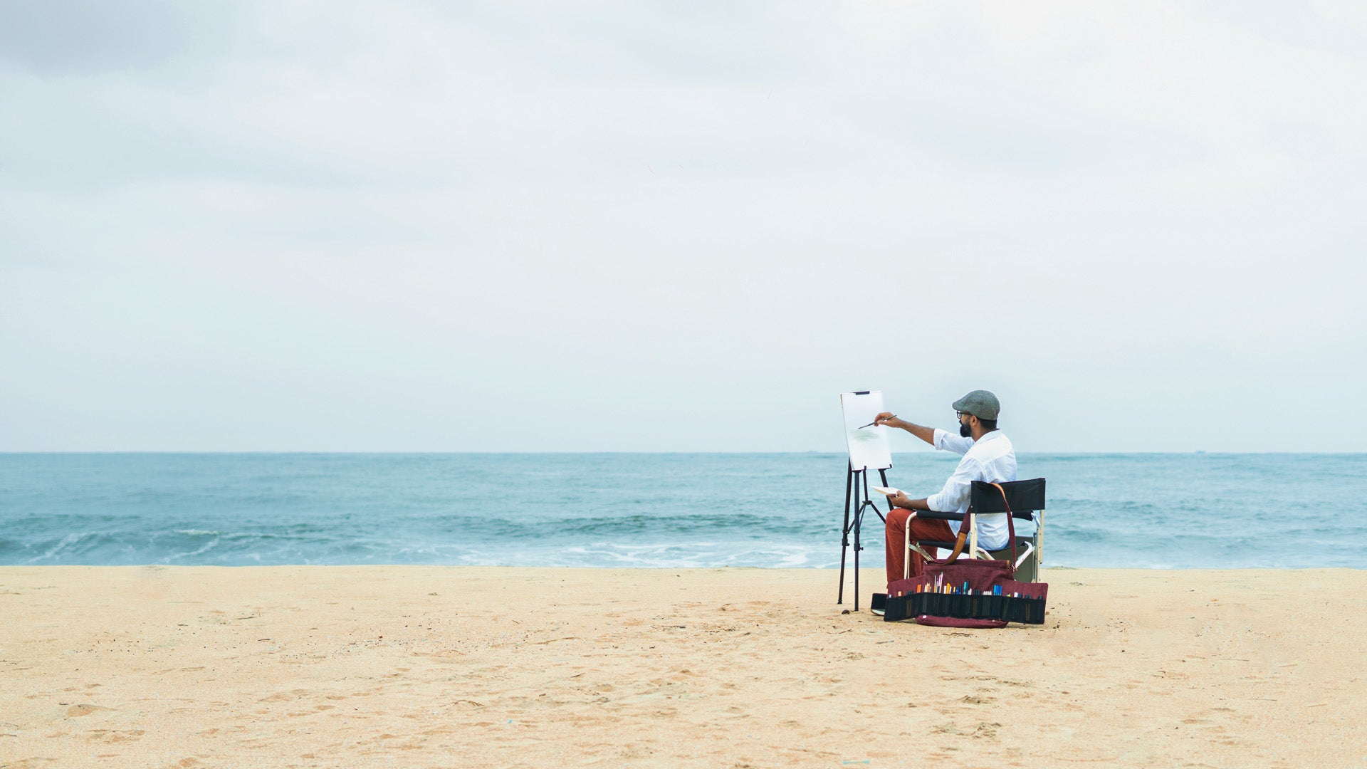 A plein-air artist painting on the beach with an open Lilorosh artist's bag beside him that neatly holds & displays all his art supplies